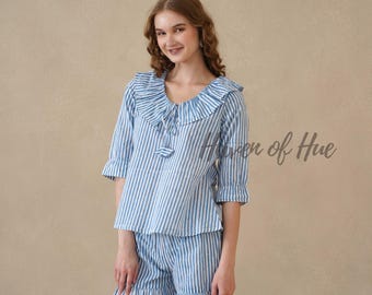 Puede incluir: Un conjunto de dos piezas compuesto por una blusa y unos pantalones cortos a rayas azules y blancas. La blusa tiene mangas tres cuartos, un cuello con volantes y cierre de lazo, y el texto Haven of Hue. El pantalón tiene un dobladillo con volantes.