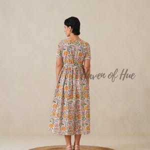 May include: A floral print midi dress in white, pink, and orange. The dress features short sleeves and a V-neckline. The text "Haven of Hue" is visible on the right side. The model is standing on a round, brown rug.