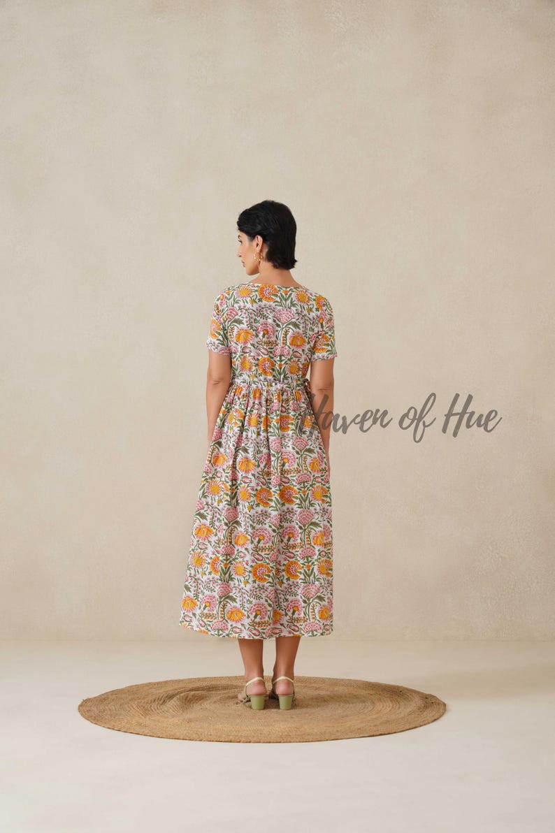 May include: A floral print midi dress in white, pink, and orange. The dress features short sleeves and a V-neckline. The text "Haven of Hue" is visible on the right side. The model is standing on a round, brown rug.