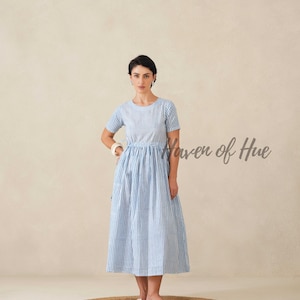 May include: A light blue and white striped dress with short sleeves. The dress has a fitted bodice and a flared skirt. The model is standing on a round, woven rug. The text "Haven of Hue" is visible on the right side of the image.