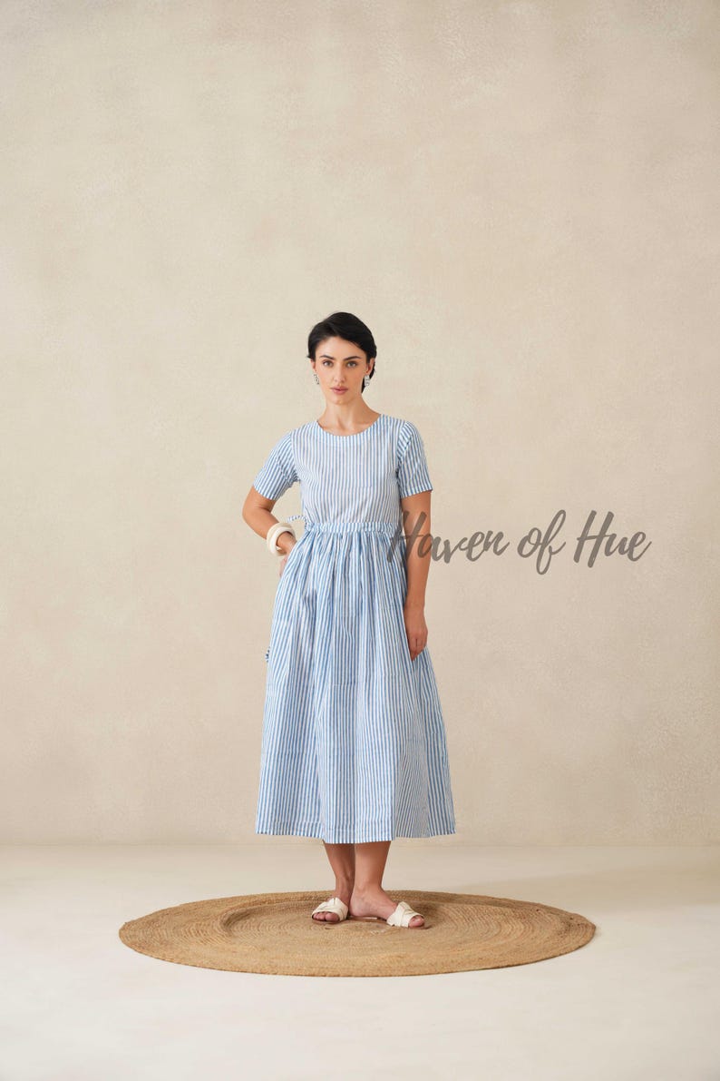 May include: A light blue and white striped dress with short sleeves. The dress has a fitted bodice and a flared skirt. The model is standing on a round, woven rug. The text "Haven of Hue" is visible on the right side of the image.