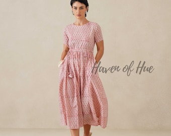 May include: A midi-length dress in a soft pink hue featuring a delicate repeating floral pattern, short sleeves, a round neckline, and a cinched waist with a drawstring tie. The text Haven of Hue is visible on the right side of the garment.