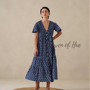 May include: A navy blue midi dress featuring a white floral pattern, a V-neckline, short flutter sleeves, and a button-down front. The dress has a tiered skirt design and includes the text Haven of Hue printed in a script font on the right side.