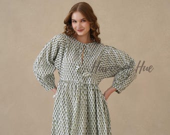 May include: A long-sleeved dress featuring a white base with a repeating green diamond-shaped pattern. The garment includes a round neckline with a central tie closure and voluminous sleeves that gather at the cuffs, creating a full silhouette.