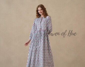 May include: A long-sleeved midi dress featuring a repeating pattern of pink tulip-like flowers on a light blue background. The dress has a gathered waist, a round neckline with a small front slit and tassel ties, and a loose, flowing skirt.