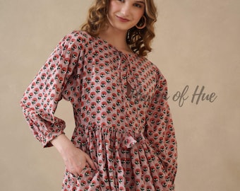 May include: A long-sleeved dress in a dusty pink hue featuring a repeating pattern of small red and green floral motifs. The garment includes a round neckline with a tie closure, gathered sleeves with cuffs, and visible side pockets at the waist.