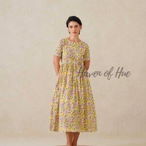 May include: A yellow floral print dress with short sleeves and a midi length. The dress features a pattern of purple flowers and green leaves. The text "Haven of Hue" is written on the right side of the dress. The dress is worn on a round, woven rug.