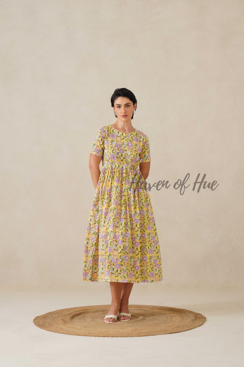 May include: A yellow floral print dress with short sleeves and a midi length. The dress features a pattern of purple flowers and green leaves. The text "Haven of Hue" is written on the right side of the dress. The dress is worn on a round, woven rug.