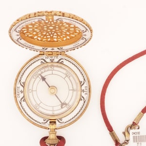 May include: A vintage-style compass pendant with a gold-colored case and a red cord. The compass face is white with black markings and a needle pointing north. The top of the compass has an openwork design. The pendant is on a white background.