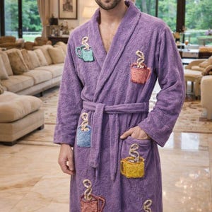 Op de afbeelding: Een zachte, lavendelkleurige badjas met geborduurde koffiekopjes. De badjas heeft een sjaalkraag, een tailleband en twee voorzakken. De koffiekopjes zijn in verschillende kleuren, waaronder blauw, roze en geel.