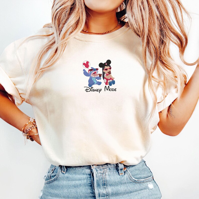 K&ouml;nnte beinhalten: Ein cremefarbenes T-Shirt mit einer kleinen Grafik auf der Brust. Die Grafik zeigt die Figur Stitch und eine stilisierte Person mit Mickey-Maus-Ohren, beide tragen eine rote Sonnenbrille. Unter den Figuren steht der Text "Disney Mode".