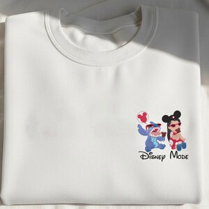 K&ouml;nnte beinhalten: Ein wei&szlig;es Sweatshirt mit Rundhalsausschnitt und einer kleinen Grafik auf der unteren rechten Seite. Die Grafik zeigt Stitch und Lilo mit roter Sonnenbrille, die braune Leckereien halten, dazu ein roter Micky-Maus-Ballon und der Text Disney Mode.