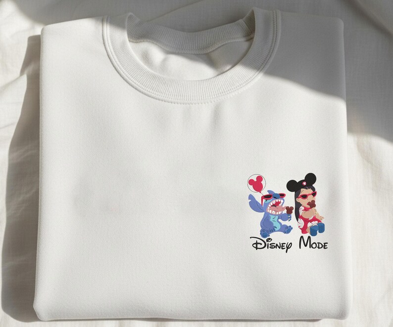 K&ouml;nnte beinhalten: Ein wei&szlig;es Sweatshirt mit Rundhalsausschnitt und einer kleinen Grafik auf der unteren rechten Seite. Die Grafik zeigt Stitch und Lilo mit roter Sonnenbrille, die braune Leckereien halten, dazu ein roter Micky-Maus-Ballon und der Text Disney Mode.
