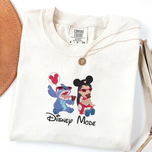 K&ouml;nnte beinhalten: Ein cremefarbenes T-Shirt mit einer Grafik von Stitch und Lilo, die rote Sonnenbrillen und Mickey-Maus-Ohren tragen. Stitch h&auml;lt einen roten Luftballon und Lilo isst eine Schokoladens&uuml;&szlig;igkeit. Der Text Disney MODE ist darunter in Schwarz gedruckt.