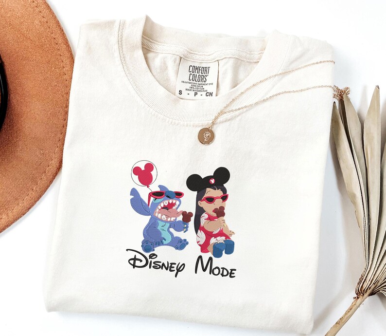K&ouml;nnte beinhalten: Ein cremefarbenes T-Shirt mit einer Grafik von Stitch und Lilo, die rote Sonnenbrillen und Mickey-Maus-Ohren tragen. Stitch h&auml;lt einen roten Luftballon und Lilo isst eine Schokoladens&uuml;&szlig;igkeit. Der Text Disney MODE ist darunter in Schwarz gedruckt.