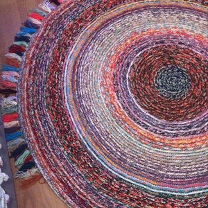 May include: A round rug with a spiral design in shades of red, purple, orange, and blue. The rug has a fringe of colorful tassels around the edge. The rug is made of woven material and is suitable for a living room or bedroom.