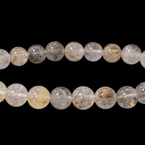 Puede incluir: Un brazalete de cuentas redondas y transparentes con inclusiones beige y grises. El brazalete está ensartado en una banda elástica y se muestra sobre un fondo negro. Las cuentas tienen aproximadamente 1 cm de diámetro.