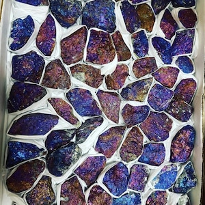 May include: A box containing many iridescent pyrite stones. The stones show a spectrum of colours, including blues, purples, and golds. Each stone is individually wrapped in white paper, creating a visually appealing arrangement.