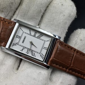Pode incluir: Um relógio de pulso retangular Seiko com mostrador branco, numerais romanos pretos e caixa prateada. O relógio possui uma pulseira de couro castanho com um padrão texturizado de imitação de crocodilo e costuras visíveis.