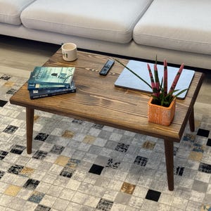 Peut inclure: Une table basse rectangulaire en bois foncé avec des livres, une tasse, une télécommande, un ordinateur portable et une petite plante en pot. La table est posée sur un tapis à motifs, près d'un canapé clair.