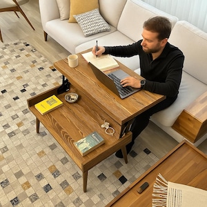 May include: A two-tiered, wooden coffee table with a lift-top design. The table is a warm brown colour and has a rectangular shape. A person is sitting on a light-coloured sofa, using a laptop and writing on a notepad. The table has a small bowl and a book on it.