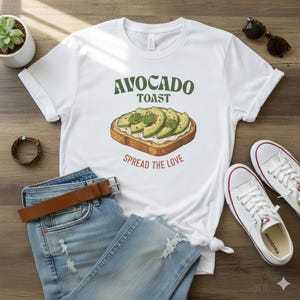 Può includere: T-shirt bianca con la scritta "AVOCADO TOAST" sopra un'illustrazione di toast con avocado. La frase "SPREAD THE LOVE" è sotto l'illustrazione. La maglietta è abbinata a jeans blu, una cintura marrone e scarpe da ginnastica bianche.