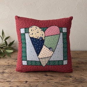 May include: A square throw pillow featuring a patchwork heart design in the center. The pillow has a red background with small black stars. The heart is composed of various fabric patterns including moons, stars, and floral prints with two buttons.