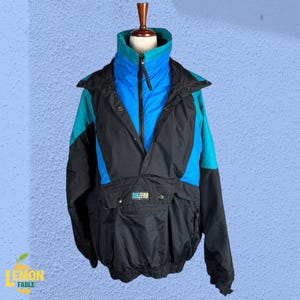 May include: A vintage anorak-style jacket in black, teal, and blue. The jacket features a high collar, a half-zip closure, and a large front pocket. The brand name "SUBZERO" is visible on the pocket. The jacket is displayed on a mannequin.