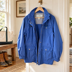 May include: A bright blue L.L.Bean jacket featuring a high collar, a full-length front zipper, and multiple pockets with snap closures. The interior lining is a light gray mesh fabric, and the jacket is displayed hanging on a wooden hanger.
