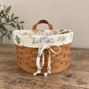 Può includere: Un cesto rotondo in legno intrecciato marrone con una fodera in tessuto color crema caratterizzata da una stampa botanica di fiori blu e foglie verdi. La fodera include una chiusura a coulisse con due lacci in tessuto e perline in legno.