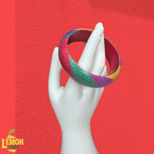 Puede incluir: Una pulsera de brazalete colorida con un patrón de piel de serpiente en tonos rosa, verde, morado, amarillo y azul. La pulsera se muestra en una mano blanca sobre un fondo rojo. El logotipo de Lemon Fable está en la esquina inferior izquierda.