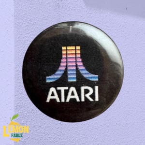 Puede incluir: Un botón redondo negro con el logotipo de Atari en un degradado de colores y la palabra "ATARI" en blanco. El botón está sobre un fondo morado claro.