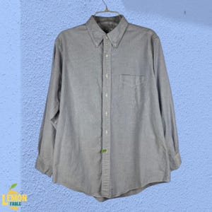Puede incluir: Camisa abotonada gris claro con mangas largas, cuello abotonado y bolsillo en el pecho. La camisa está colgada de una percha metálica sobre un fondo azul claro texturizado. La camisa tiene un estilo clásico e informal.