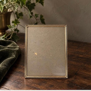 May include: A rectangular picture frame with a thin, metallic gold-toned border featuring a subtle textured pattern along the edges. The frame contains a plain, light brown backing material with small, dark speckles visible throughout the surface.