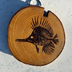 May include: A round, wooden slice with a dark brown, burnt-wood fish skeleton design. The fish has a long snout and spiky fins. A silver metal ring is attached to the top for hanging. The wood has visible growth rings.