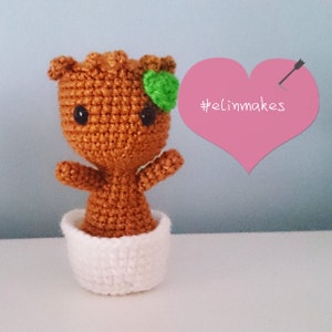 Pode incluir: Figura amigurumi feita à mão, representando uma personagem de filme, em fio castanho com um detalhe de folha verde. Está num vaso branco de crochê, com olhos pretos. O texto "#elinmakes" está num coração rosa.