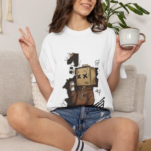 Può includere: Una t-shirt bianca a maniche corte con una stampa grafica di un personaggio con una testa a scatola di cartone marrone, che indossa una felpa marrone con cappuccio e tiene in mano una tazza di caffè. La testa a scatola ha croci nere per occhi e una bocca a linea semplice, circondata da forme e linee astratte a inchiostro nero.