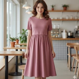 May include: A dusty rose-colored dress with short sleeves and a gathered waist. The dress has a round neckline and a flared skirt, creating a classic silhouette. The woman is standing in a cafe setting with wooden tables and chairs.