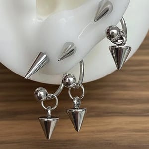 May include: A set of silver-toned metal body jewelry featuring multiple spike and ball attachments. The design includes two circular hoops with dangling cone-shaped spikes and two stationary spikes attached directly to the curved metal base.