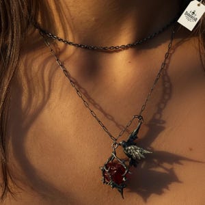 Op de afbeelding: Een ketting met twee strengen en een donkerzilveren afwerking. De langere ketting heeft een hanger met een vogel die boven een kooi met een rode steen zit. Het label luidt "DARKWEAR JEWELRY CO."