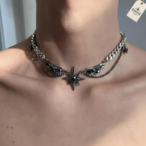 Könnte beinhalten: Silberfarbene Halskette mit Sternanhänger und Flügelakzenten. Die Halskette hat mehrere Kettenschichten und ein Etikett mit der Aufschrift "DARKWEAR JEWELRY CO."