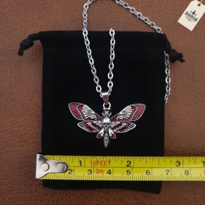 Puede incluir: Un collar con colgante de polilla plateado con un detalle de calavera en el tórax y detalles en esmalte rojo en las alas. Una cinta métrica amarilla muestra un ancho de unos 5 cm. El collar se presenta sobre una bolsa negra.