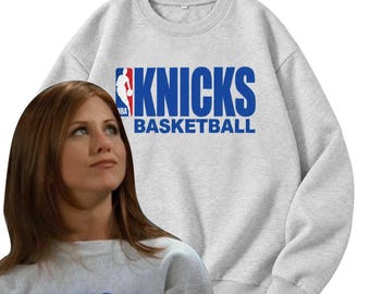 Könnte beinhalten: Ein hellgraues Sweatshirt mit Rundhalsausschnitt, auf dessen Brust der blaue Schriftzug KNICKS BASKETBALL sowie das rot-weiß-blaue NBA-Logo aufgedruckt sind. Das Kleidungsstück hat lange Ärmel, einen gerippten Halsausschnitt und gerippte Bündchen.