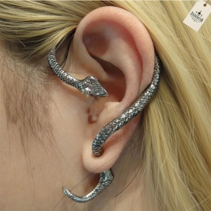 May include: A silver-coloured ear cuff shaped like a snake, coiled around the ear. The snake's head is near the top of the ear, with the body curving downwards. The jewellery has a textured, scale-like pattern. The tag says DARKWEAR JEWELRY.