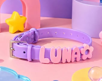 May include: A light purple silicone collar featuring the name LUNA in raised pink block letters and a pink flower charm. The collar includes a silver-toned metal buckle and a series of adjustment holes, designed for a pet to wear around the neck.