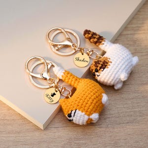 May include: Two crochet cat keychains with gold-colored hardware. One cat is orange and white, the other is white and brown. Each keychain has a small, round charm with a cat silhouette and a name: "Kitty" and "Shadow".