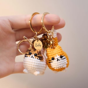 May include: Two crochet cat keychains with gold-colored hardware. One cat is white, brown, and tan, and the other is orange and white. Each keychain has a gold-colored ring and a small gold-colored tag that says "Baby".