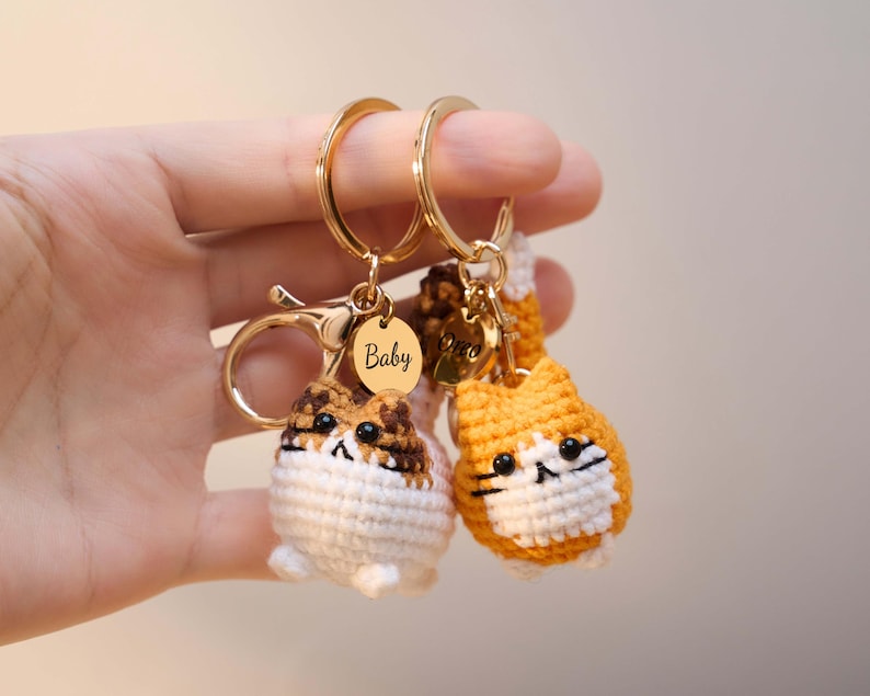 May include: Two crochet cat keychains with gold-colored hardware. One cat is white, brown, and tan, and the other is orange and white. Each keychain has a gold-colored ring and a small gold-colored tag that says "Baby".