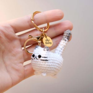 May include: A handmade crochet cat keychain in white and gray. The cat has black eyes and whiskers, with a gold-colored metal clasp and a small charm that says "Baby". The keychain is held in a hand.