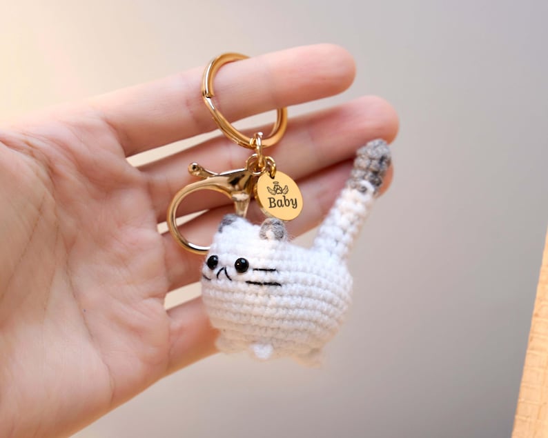 May include: A handmade crochet cat keychain in white and gray. The cat has black eyes and whiskers, with a gold-colored metal clasp and a small charm that says "Baby". The keychain is held in a hand.